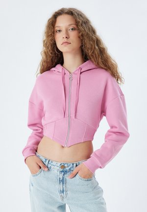 Rosa, kurz geschnittenes Hoodie mit langen Ärmeln, Reißverschluss vorne und Kapuze. Ist aus einem strukturierten Stoff gefertigt und hat eine figurnahe Silhouette. Getragen mit hellblauen Jeans.