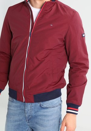 Mężczyzna ubrany w bordową kurtkę bomberkę z granatowymi mankietami i ściągaczem, częściowo rozpiętą na białą koszulę i jasnoniebieskie dżinsy.