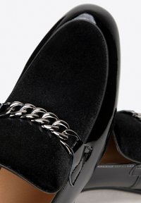 Zwarte loafers met een glanzende patenten leren afwerking en een zachte fluwelen bovenkant, versierd met een zilveren kettingdetail over de voorkant.