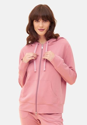 Giovane donna che indossa una felpa con cappuccio rosa con zip e pantaloni abbinati, tenendo i lacci del cappuccio con entrambe le mani, su uno sfondo semplice.