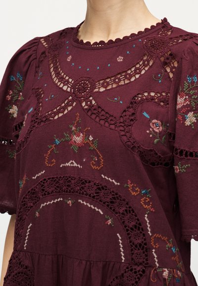 Μπορντό μπλούζα με κεντήματα με λουλούδια, δαντελωτές περικοπές, φουσκωτά μανίκια και περίπλοκα σχέδια κροσέ στο στήθος και στους ώμους.