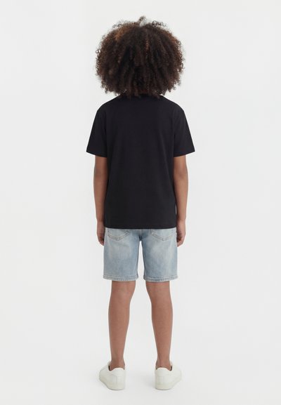 Παιδί με σγουρά μαλλιά που φοράει μαύρο μπλουζάκι, ανοιχτόχρωμο τζιν σορτς και λευκά αθλητικά παπούτσια, στέκεται με την πλάτη γυρισμένη σε λευκό φόντο.