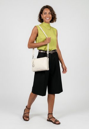 Mujer sonriendo, con top sin mangas de color verde lima, pantalones cortos negros hasta la rodilla, sandalias marrones y llevando un bolso blanco cruzado al hombro.