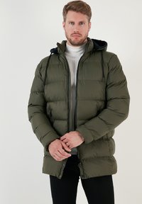 Maslinasto zelena puffer jakna sa patentnim zatvaračem i dugmetom, podesivim kapuljačom i dva bočna džepa; karakterizira je kvadratna tekstura i crni detalji.