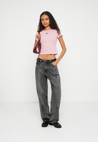 Ροζ cropped t-shirt με μπλε λογότυπο, συνδυασμένο με χαλαρά μαύρα τζιν και μαύρα loafers. Κρατώντας μια μικρή μπορντό τσάντα χειρός.