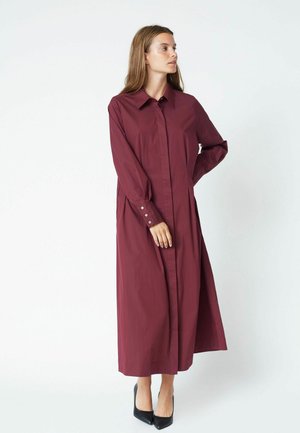 Abito camicia lungo borgogna realizzato in tessuto morbido, con colletto, maniche lunghe e polsini abbottonati. Indossato con tacchi neri.