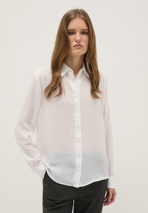 Junge Frau mit langem Haar, die eine durchsichtige weiße Bluse mit Knöpfen und schwarze Hose trägt und vor einem schlichten hellen Hintergrund steht.