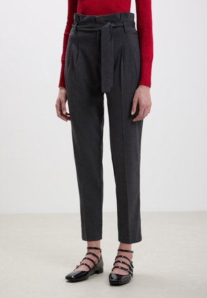 Pantaloni grigi su misura con vita alta e cintura in tessuto, caratterizzati da pieghe laterali e gambe affusolate. Abbinati a sandali neri con cinturini.