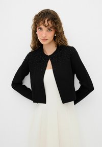 Cardigan nero cropped realizzato in tessuto knit, con una decorazione di perline lungo il colletto e maniche lunghe. Indossato sopra un vestito bianco.