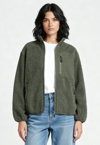 Donna che indossa una giacca in pile verde oliva con tasche sul petto e sui lati con zip, camicia bianca e jeans blu, in piedi davanti a uno sfondo semplice.
