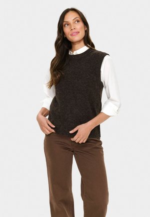 Frau in einem dunkelbraunen ärmellosen Pullover über einem weißen Hemd und braunen Hosen, die mit den Händen in der Taille steht und leicht nach oben und rechts schaut.