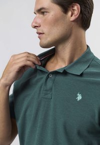 Tumši zaļa polo krekls no teksturētas auduma, ar klasisku apkakli un mazu izšūtu logotipu kreisajā krūtī.