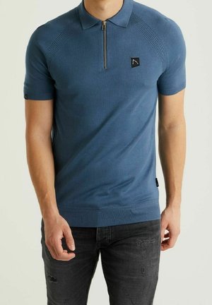 Niebieska koszulka polo z krótkim rękawem i suwakiem przy kołnierzu, z prążkowaną fakturą, naszywką z logo na piersi oraz dopasowanym krojem. Noszona z czarnymi, przetartymi dżinsami.