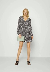 Mulher de pé, vestindo um vestido de mangas longas com padrão preto e branco, sapatos de salto aberto pretos, segurando uma bolsa verde clara contra um fundo neutro.