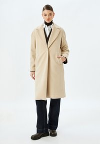 Beige uldfrakke med notch-krave, enkelt knaplukning, sidelommer og lige pasform, parret med mørke jeans og sorte støvler.