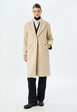 Beige wollen jas met een geknikte kraag, enkele knoopsluiting, zijzakken en een rechte snit, gecombineerd met donkere jeans en zwarte laarzen.