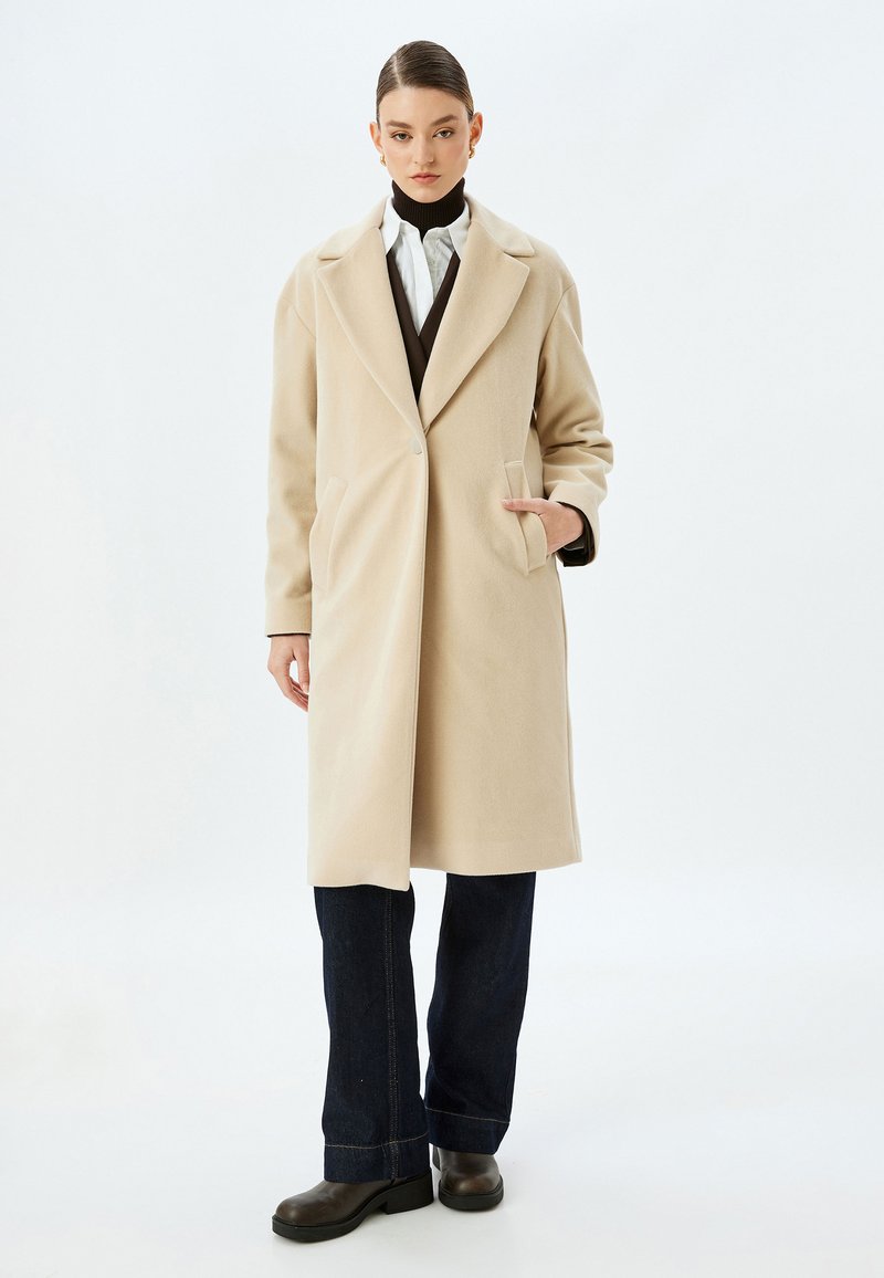Beige uldfrakke med notch-krave, enkelt knaplukning, sidelommer og lige pasform, parret med mørke jeans og sorte støvler.