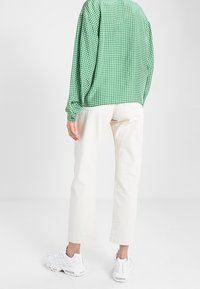 Chemise à manches longues à carreaux verts, coupe ample, associée à un pantalon droit de couleur crème. Des baskets blanches complètent la tenue.