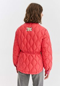 Žena viděná zezadu, která má na sobě korálově zbarvenou prošívanou bundu s diamantovým stehem a stahovací šňůrkou v pase, v kombinaci se šedými kalhotami na bílém pozadí.