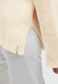 Beige, fuzzy sweater with a high-low hem and side slits, paired with white pants featuring a patch pocket and label on the hem.