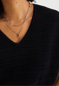 Černý žebrovaný top s výstřihem do V; vrstvené zlaté náhrdelníky s různými styly řetězů a obdélníkovým přívěskem. Detaily texturované látky.