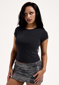 Μαύρο εφαρμοστό τοπ με κοντά μανίκια και γκλίτερ κείμενο «Strong». Συνδυάζεται με ασημένια μίνι φούστα από παγιέτες με ρυτίδες και κάθετες γραμμές.