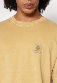 Camisa de algodón de manga corta y cuello redondo en color amarillo desgastado, con una etiqueta cuadrada con el logo de Carhartt en naranja en el lado izquierdo del pecho.