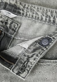 Tissu en denim gris présentant une texture délavée, une doublure blanche contrastante et une fermeture éclair argentée. Comprend une fermeture à bouton et des coutures visibles.