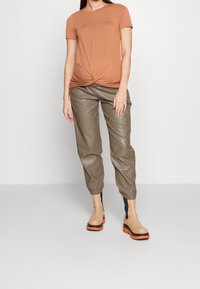 Brown knotted t-shirt, olive green leather joggers, and ankle boots with beige and orange accents. Smooth fabric and structured fit.