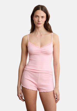 Vrouw draagt een lichtroze geribbelde top met kantafwerking en bijpassende korte broek, staand tegen een effen witte achtergrond.