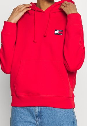Osoba si upravuje jasně červenou mikinu Tommy Jeans s předním kapsou, v kombinaci s modrými džínami, na jednoduchém světlém pozadí.