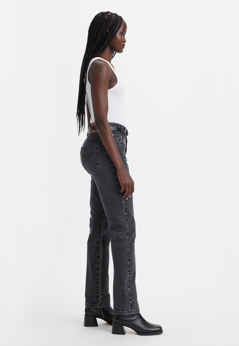 Jeans en denim gris à coupe droite, avec des accents de boutons sur les côtés et une taille haute, associés à un débardeur blanc et des bottes à talons noires.