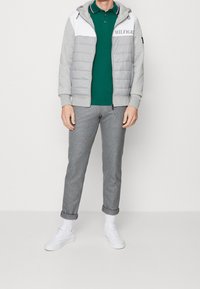 Grijze pufferjack met witte panelen en logo, groene polo, grijze geruite broek en witte sneakers. Casual outfit.