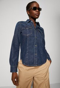 Denim srajca v temno modri barvi s ovratnikom, dvema žepoma na prsih in gumbi za zapiranje. Dolgi rokavi z gumbnimi manšeti.
