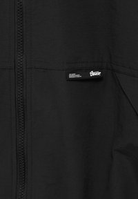 Section de veste noire avec une fermeture éclair verticale à gauche et une petite étiquette affichant « STAY UNITED » ainsi que des coordonnées à droite.