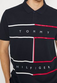 Mustat polo-paita, jossa on kontrastivärinen graafinen kuvio, jossa on punaisia ja valkoisia viivoja. "TOMMY HILFIGER" on painettu valkoisena keskelle.