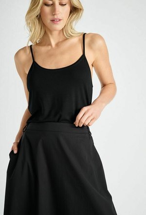 Blonde vrouw die een zwarte spaghetti top en een bijpassende zwarte rok draagt, met één hand in de zak van de rok.