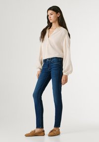 Νεαρή γυναίκα όρθια, φοράει μπλούζα με μακριά μανίκια σε εκρού χρώμα, σκούρο μπλε στενά τζιν και ταμπά μοκασίνια, μπροστά σε απλό φόντο.