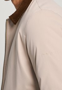 Lyse beige jakke med en tekstureret brun corduroy-krave. Diskret branding præget på ærmet, med en glat tekstilfinish.