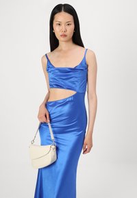 Vestido azul de cetim com um topo cortado, detalhe recortado e uma saia justa. A modelo segura uma bolsa bege clara com uma alça de ombro.