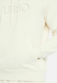 Creme hoodie med en frontlomme, snøre i hætten og prægede logo. Glat tekstur med ribbede ærmer og kant. Subtile stikninger.