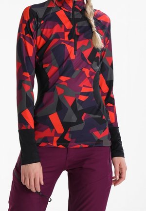 Obcisła bluzka z długim rękawem o geometrycznym wzorze w kolorach czerwonym, czarnym i fioletowym, z kołnierzem na zamek, noszona z fioletowymi spodniami, dłoń spoczywa na udzie.