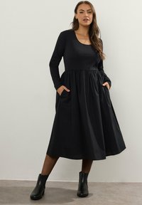 Nainen, jolla on päällään pitkähihaisen mustan midi-mekon taskut ja mustat nilkkasukat, seisoo vaaleaa seinää vasten.