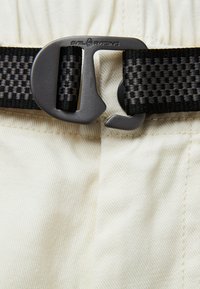 Ljust beige tyg med en texturerad yta, prytt med ett svart vävt bälte och en matt grå spänne märkt "Sail Racing."