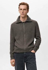 Suéter gris acanalado con cierre de cremallera y cuello, con mangas largas y diseño ajustado, combinado con vaqueros oscuros. Textura suave.