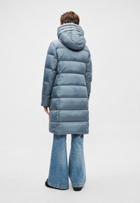 Blauwe pufferjas met capuchon, met doorgestikte gedeeltes. De jas loopt door tot halverwege de dij en is gecombineerd met wijde blauwe spijkerbroeken.