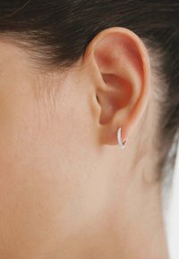 Pendiente de aro de plata con un diseño delgado, que presenta una fila de pequeñas piedras transparentes a lo largo de un borde, sujeto a una oreja perforada.