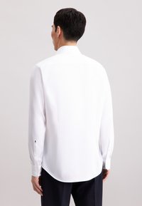 Camicia bianca a maniche lunghe con tessuto texturizzato, colletto a punta e orlo curvo. Presenta polsini con bottoni e dettagli a contrasto neri.