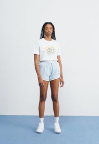 Young woman with long braids stands wearing white floral graphic t-shirt, light blue drawstring shorts, white socks, and white sneakers.