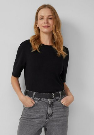 Femme aux longs cheveux blonds portant un haut noir à manches courtes rentré dans un jean gris clair taille haute avec une ceinture noire, les mains dans les poches.